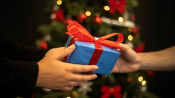 A closeup of a wrapped present being given with a christmas tree in the background.