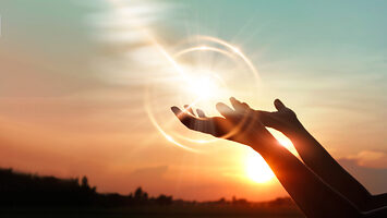 A person's hands reach, palm upward, toward the sky. A ball of light cast from the setting sun sits just above them.