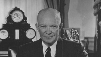 President Eisenhower behind his desk in the Oval Office, holding some papers, with his eyeglasses resting in front of him.