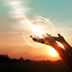 A person's hands reach, palm upward, toward the sky. A ball of light cast from the setting sun sits just above them.