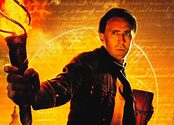 Nicolas Cage holds a torch and a book while standing in front of floating text and several large national monuments.
