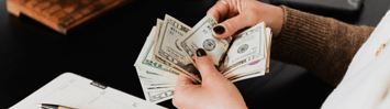 A person counts money while sitting at their desk. A pad of paper, a pen, and a phone are on the desk. 