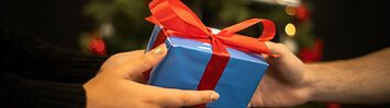 A closeup of a wrapped present being given with a christmas tree in the background.