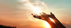 A person's hands reach, palm upward, toward the sky. A ball of light cast from the setting sun sits just above them.